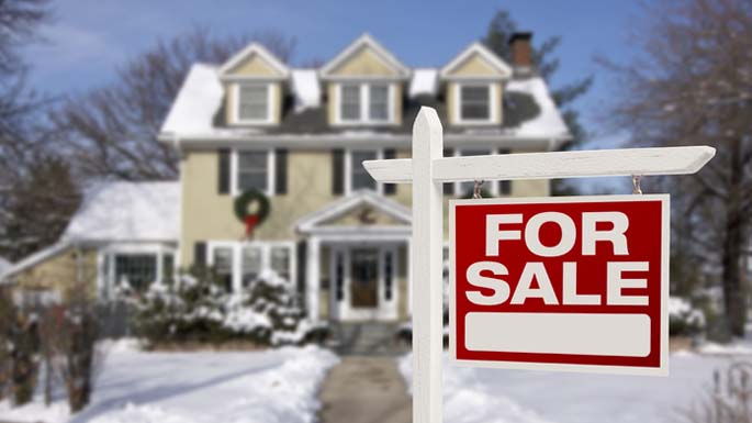 A For Sale sign in the front yard of a snow-covered home.